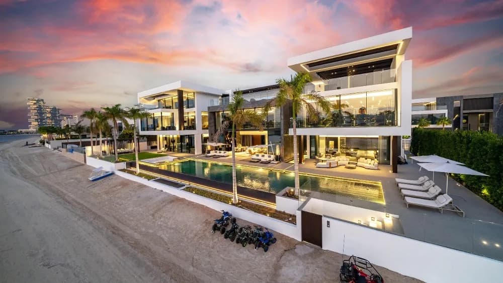 A beachfront villa at dusk, its pool lit, palms along the sand